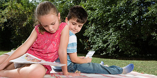 Mädchen mit Zeitschrift und Junge mit Handspielgerät auf Wiese